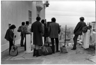 Group of hippies with bags, sleeping bags, and guitar waiting for a ride in San Francisco